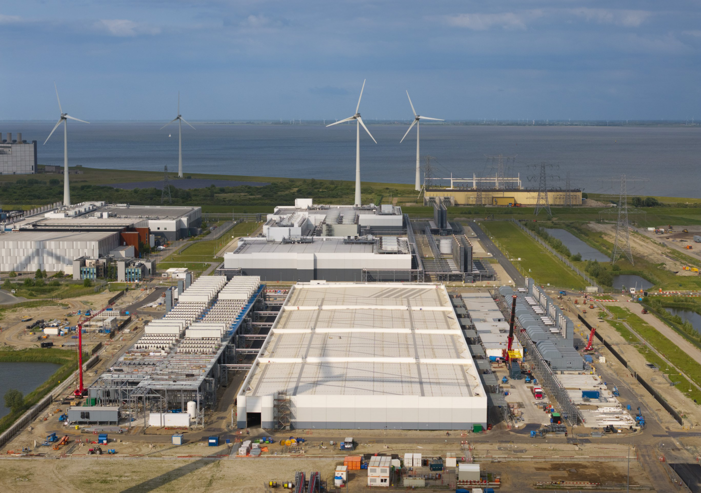 Aerial view of a large data center facility under construction, featuring multiple wind turbines and coastal energy infrastructure in the background.
