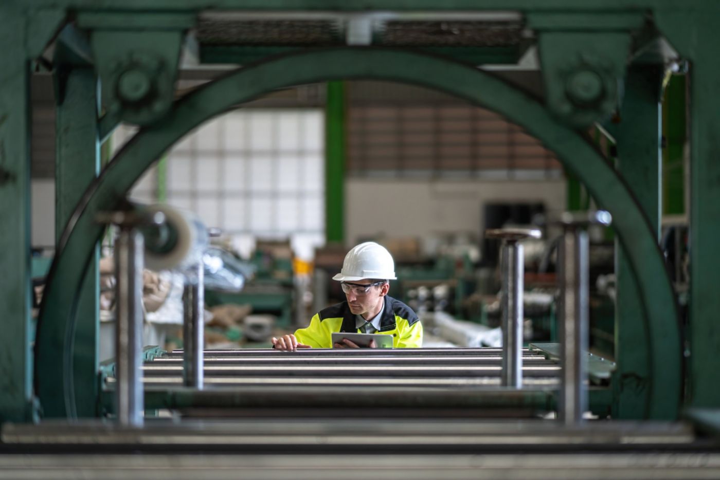 An engineer in protective gear reviews data on a tablet while inspecting machinery within a factory setting.