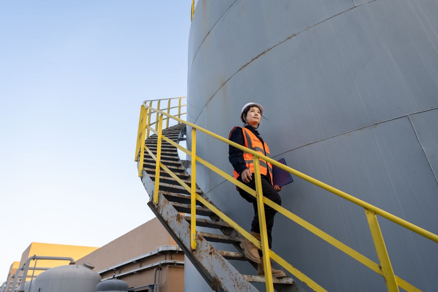 A female industrial engineer wearing a hard hat and safety vest walks down external stairs of a large storage tank while holding a blue clipboard.
