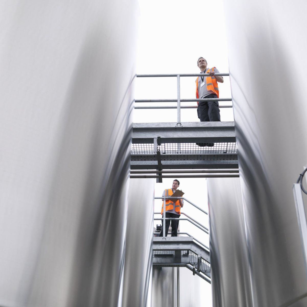 Two workers in high-visibility vests conduct a safety inspection while standing on a gantry between large stainless steel industrial storage tanks.