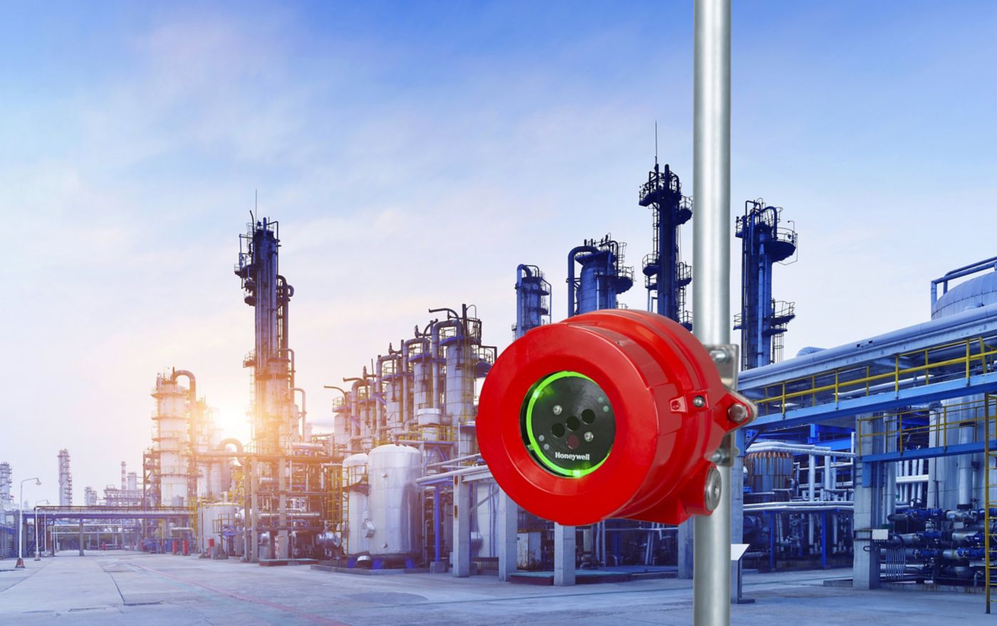 A bright red Honeywell gas detection sensor mounted on a pole against the backdrop of a petrochemical refinery facility at sunset.