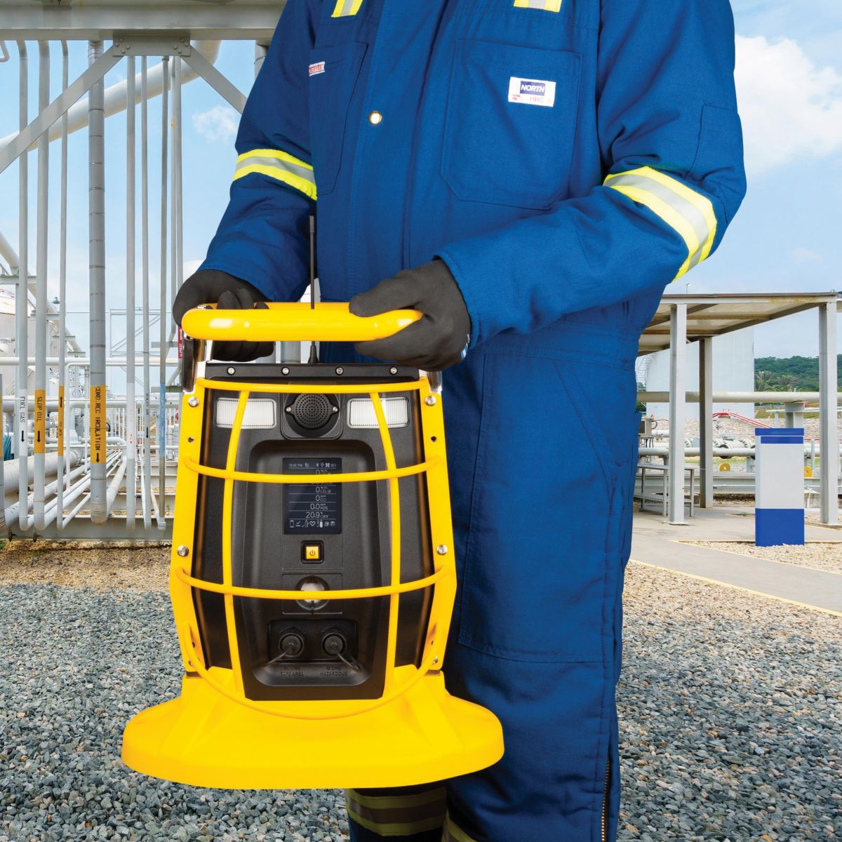 An industrial worker holds a portable gas detection monitor while standing at a processing facility featuring outdoor pipeline infrastructure.
