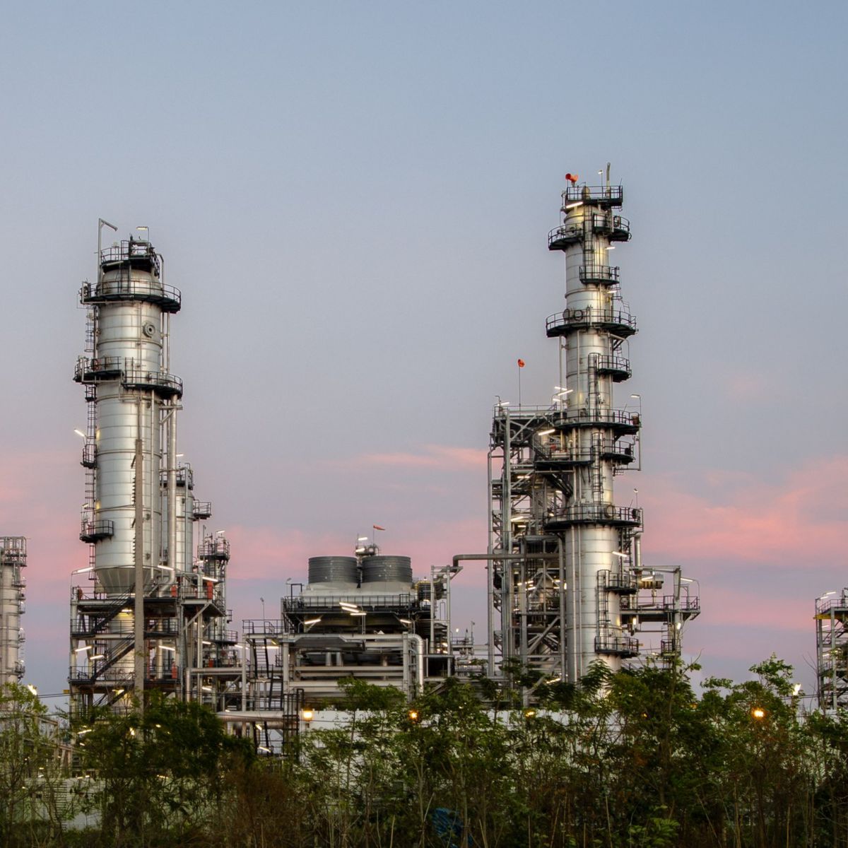 Illuminated metal distillation towers of a petrochemical facility stand against a soft sunset sky.