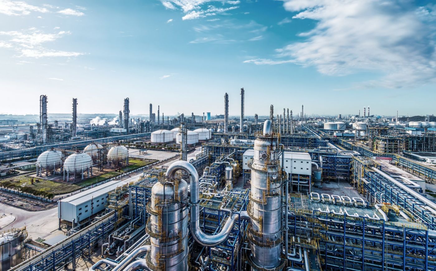 An aerial view captures the vast infrastructure of a sprawling petrochemical refinery complex under a bright, clear blue sky.
