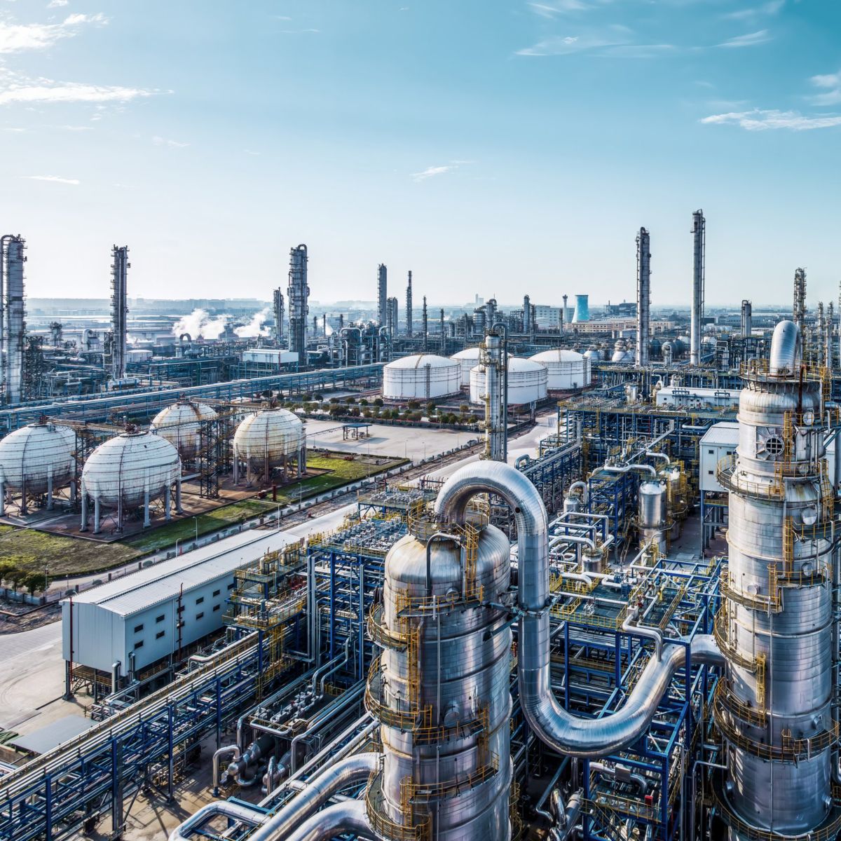An aerial view captures the vast infrastructure of a sprawling petrochemical refinery complex under a bright, clear blue sky.