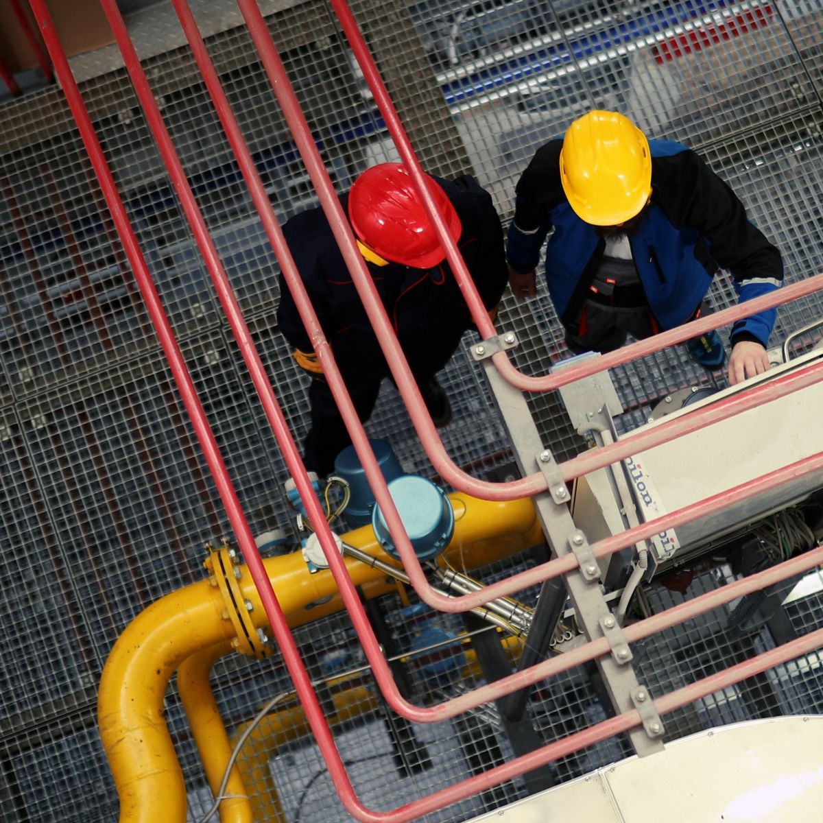 Two technicians wearing safety helmets inspect mechanical equipment and piping in an industrial facility from an overhead perspective.