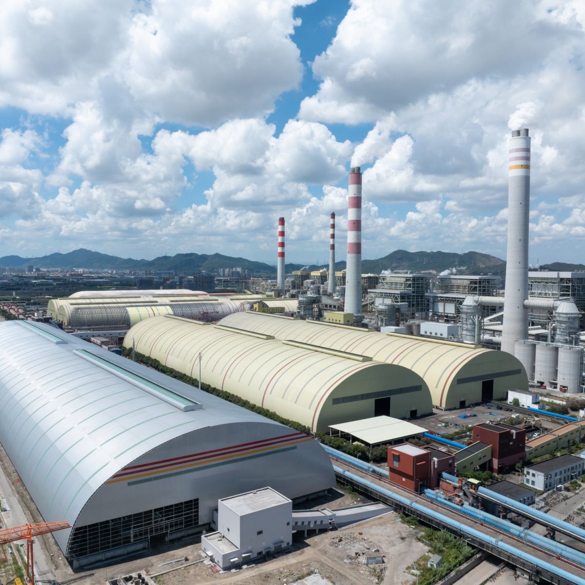 Aerial view of a large-scale power plant complex featuring storage warehouses and towering smokestacks under a cloudy blue sky.