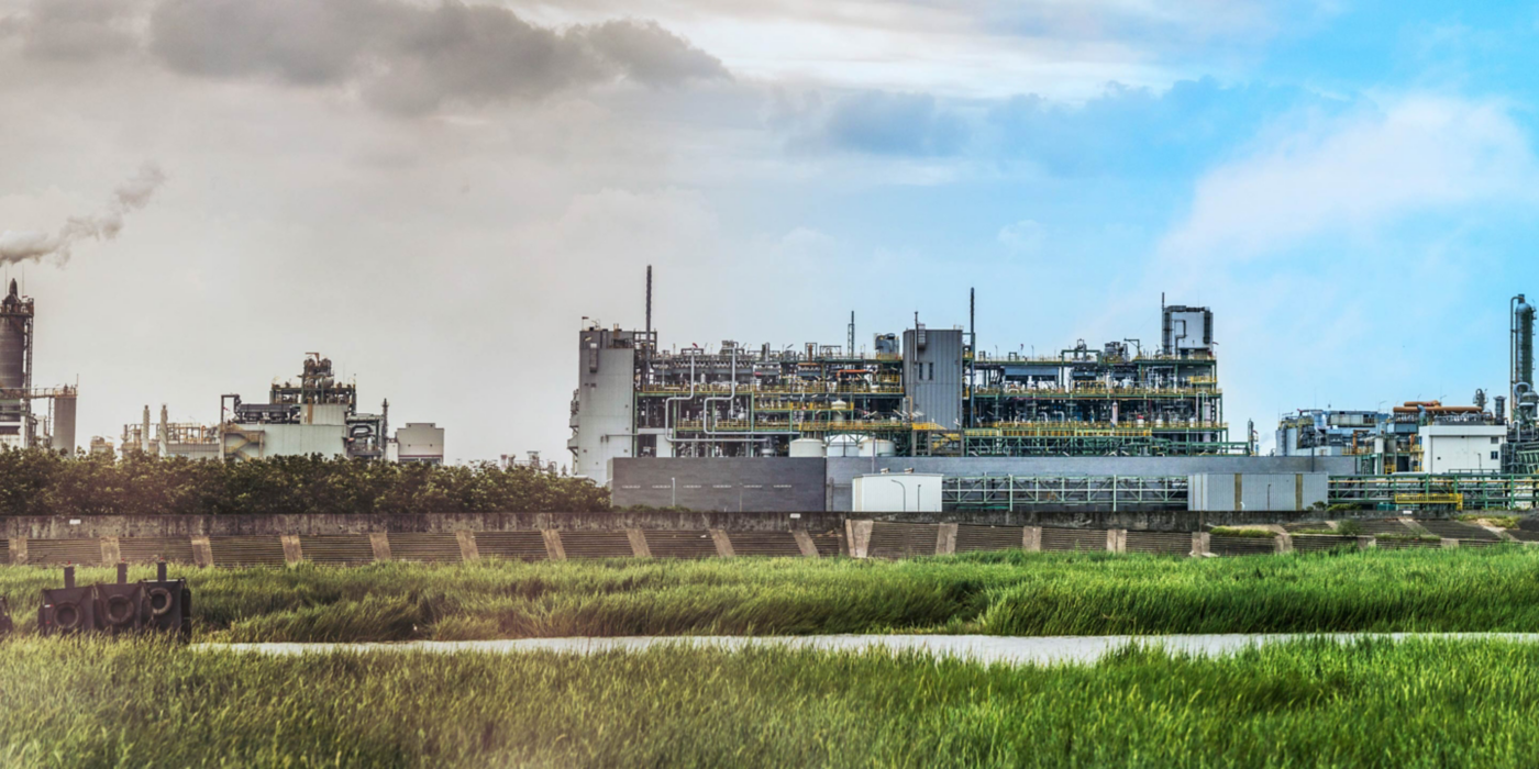 Large industrial processing facility surrounded by green fields and infrastructure under a clear sky