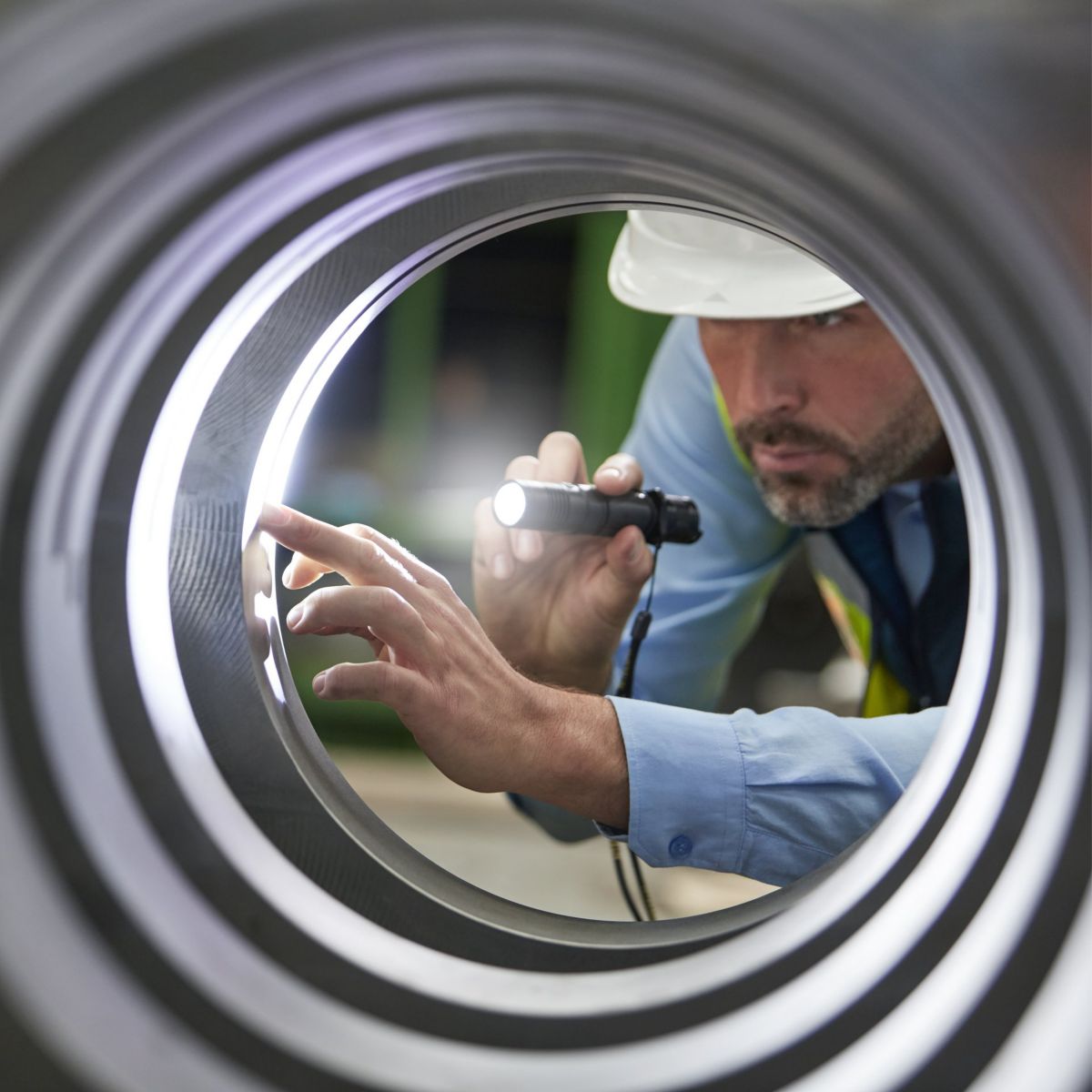A technician inspects a precision-machined metal component for defects using a flashlight, viewed through the circular frame of the industrial part.