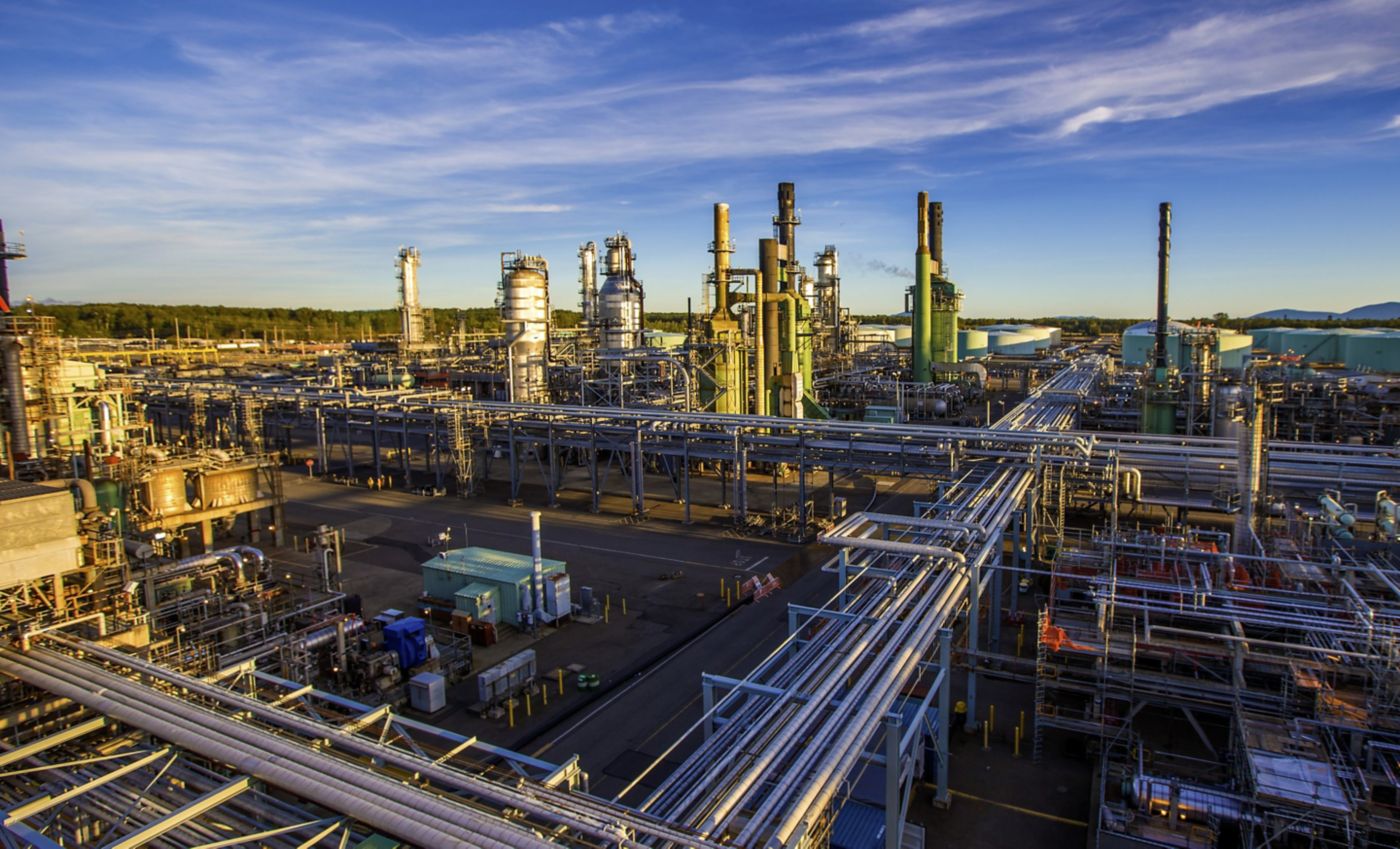 A wide-angle view of a sprawling industrial refinery complex with extensive piping systems and towering processing units under a clear blue sky.