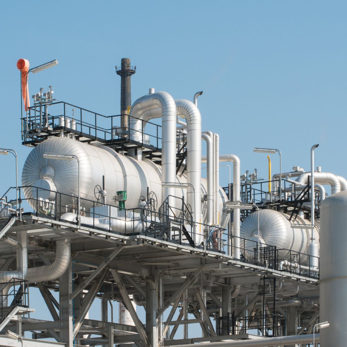 A detailed view of high-pressure storage tanks, complex piping systems, and steel gantries within an industrial oil refinery against a clear blue sky.