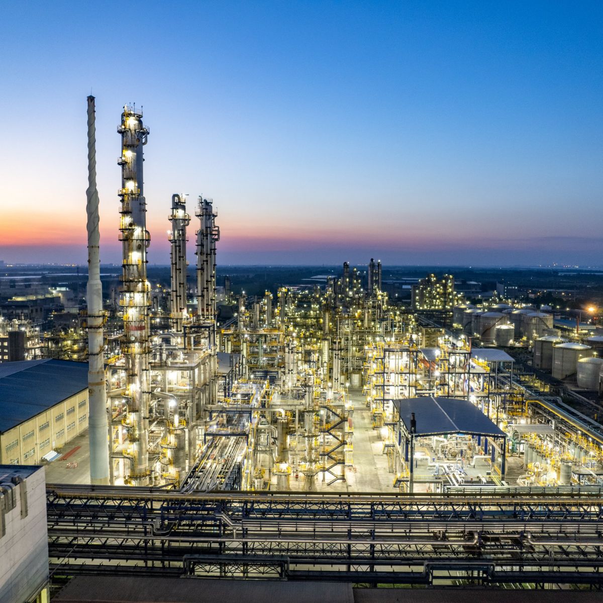 Aerial evening view of a sprawling industrial refinery complex illuminated by artificial lights against a vibrant gradient sunset sky.