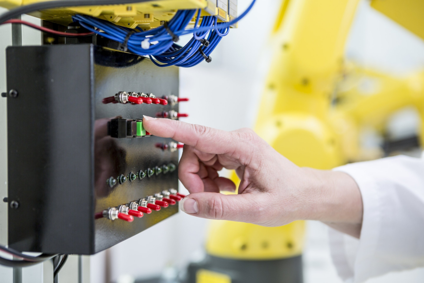 A technician presses a green button on a control panel to operate a yellow industrial robotic arm in a manufacturing facility.