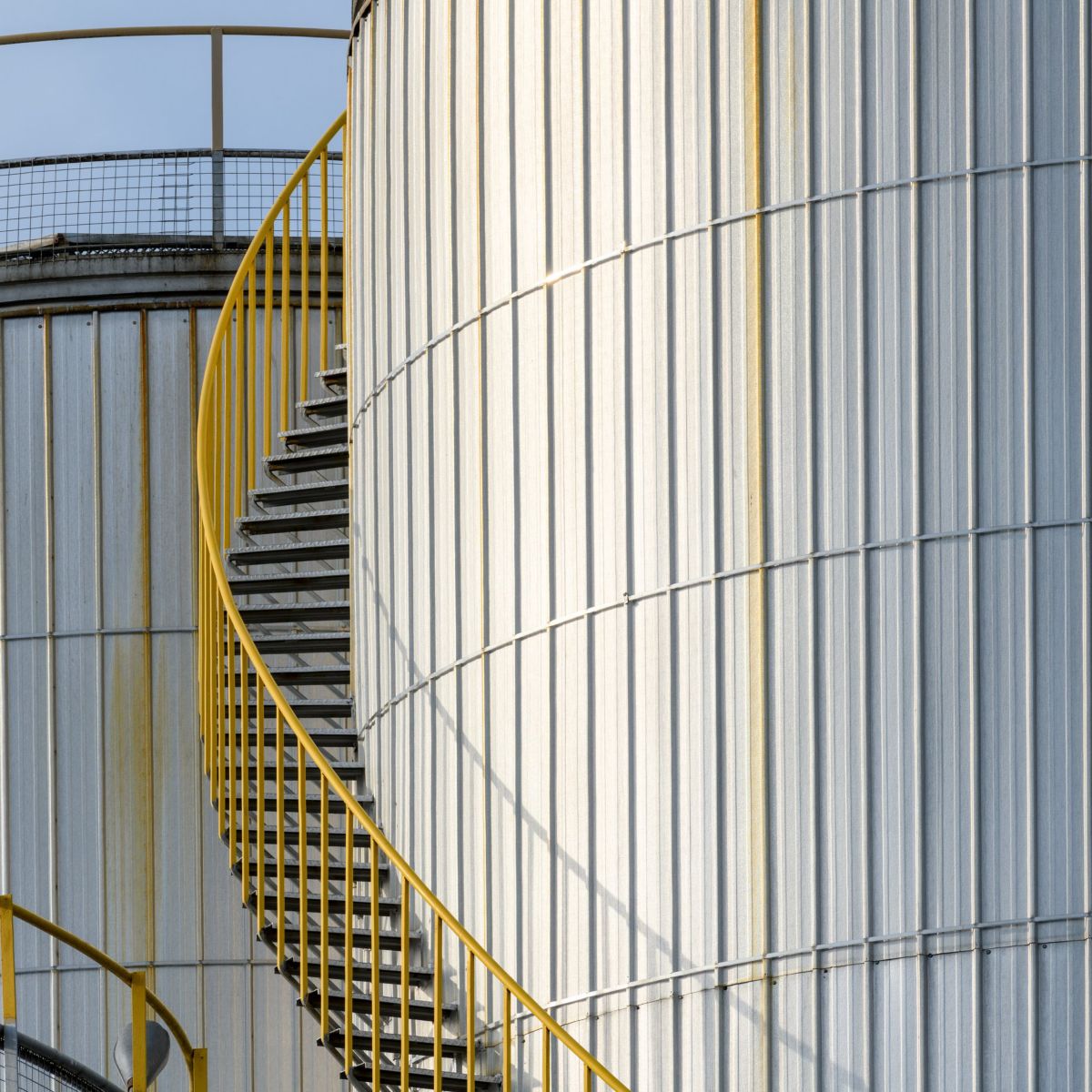 A bright yellow spiral staircase wraps around the corrugated metal exterior of large industrial storage tanks against a clear sky.