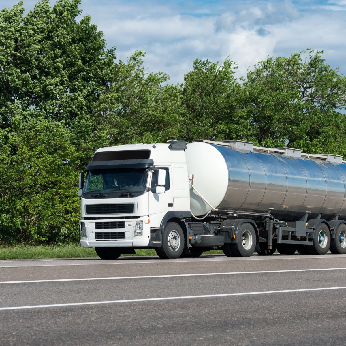 A white semi-truck with a stainless steel tanker trailer drives along a scenic highway lined with lush green trees under a partly cloudy sky.