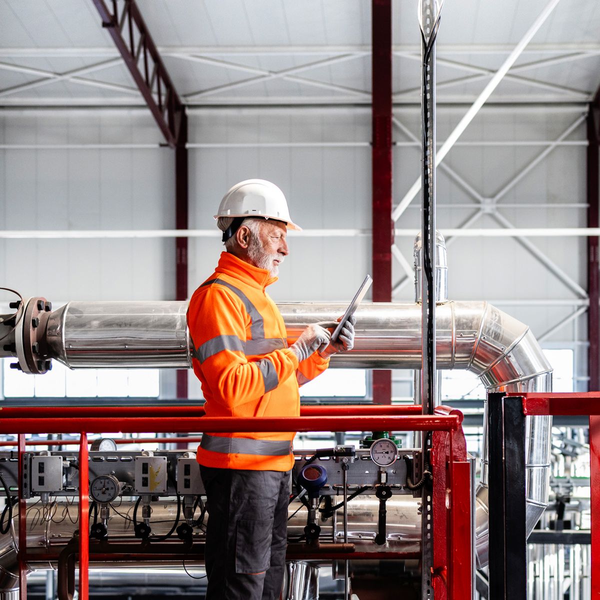 Professional in safety gear reviewing equipment data on a tablet within an industrial plant, emphasising safety compliance, monitoring, and maintenance operations.