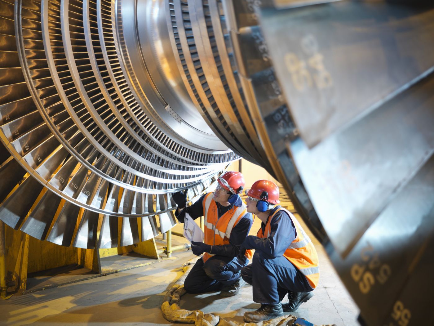Technicians wearing safety gear inspect a large power generation turbine rotor in an industrial facility.