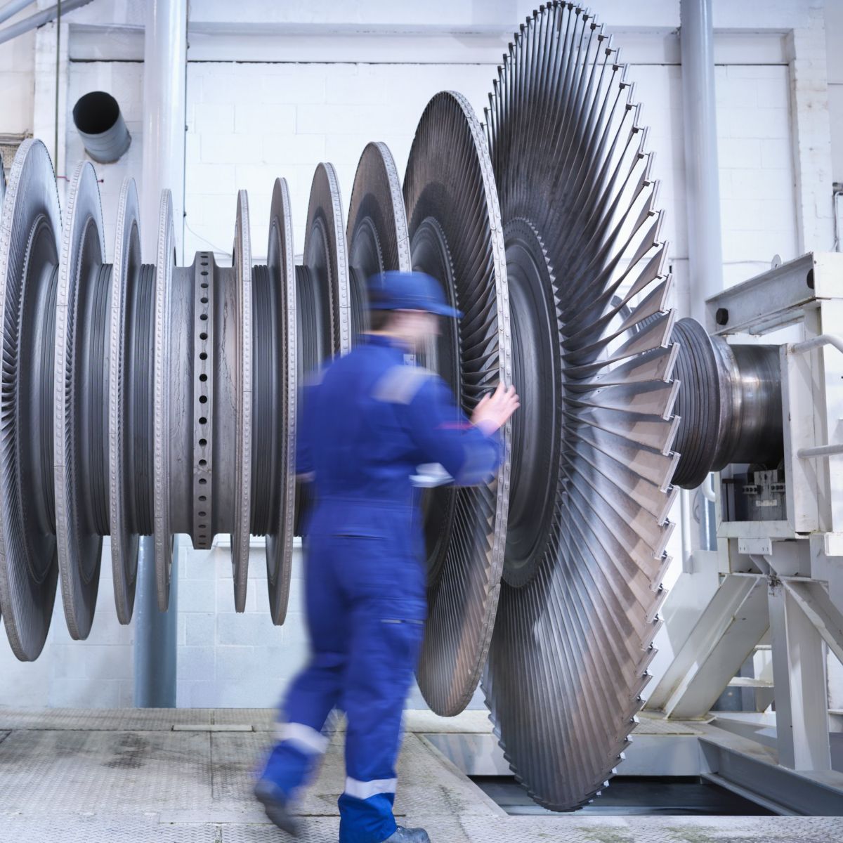 A technician in blue workwear inspects a large industrial turbine rotor within a manufacturing facility.