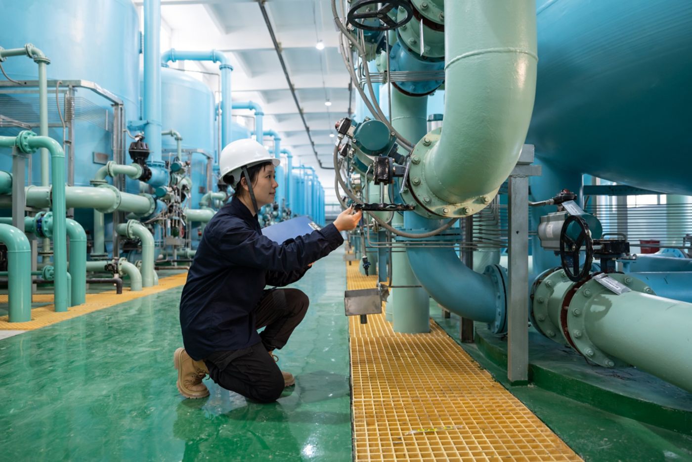 A female technician in protective gear inspects industrial piping and valves inside a modern water treatment facility.