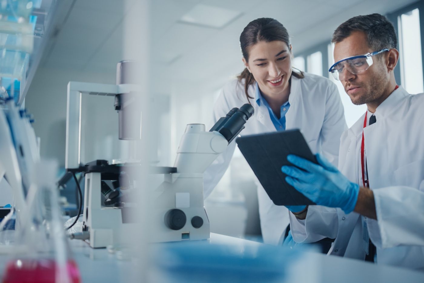 Modern Medical Research Laboratory: Portrait of Two Scientists Working, Using Digital Tablet, Analyzing Samples, Talking. Advanced Scientific Pharmaceutical Lab for Medicine, Biotechnology Development
