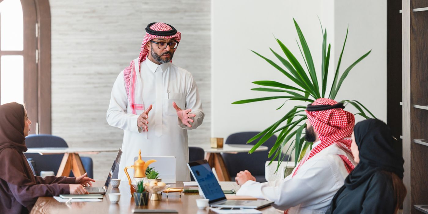 Group seated around a table during a business meeting with laptops while one participant presents to the team.
