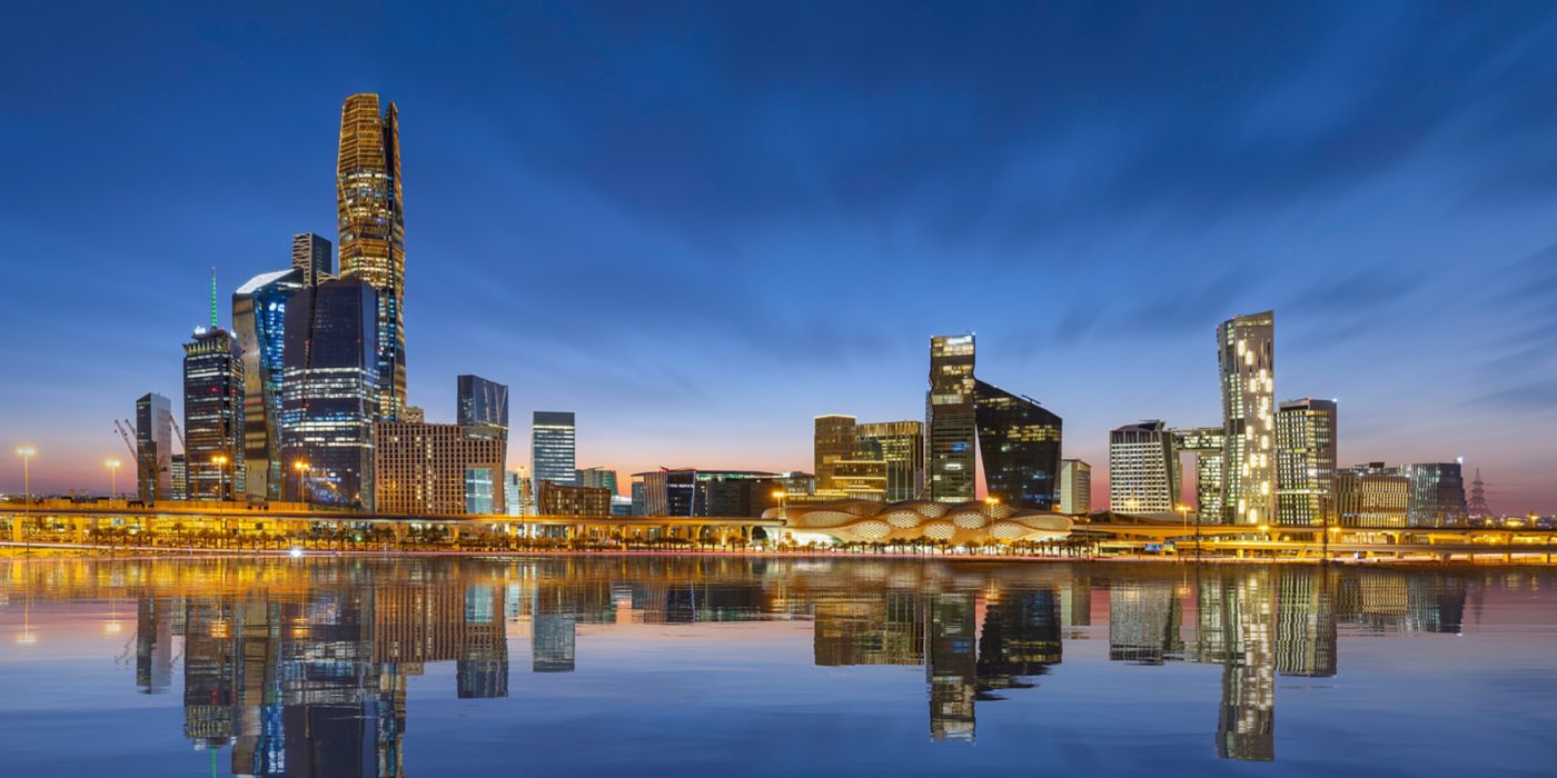 City skyline with high-rise buildings reflected in calm water, featuring illuminated architecture in an urban environment.
