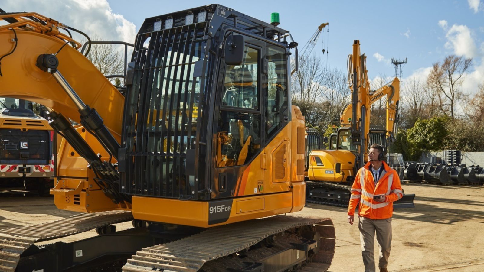 Tracked excavator with enclosed cab on a work site, with a worker in high-visibility vest and additional machinery in the background.