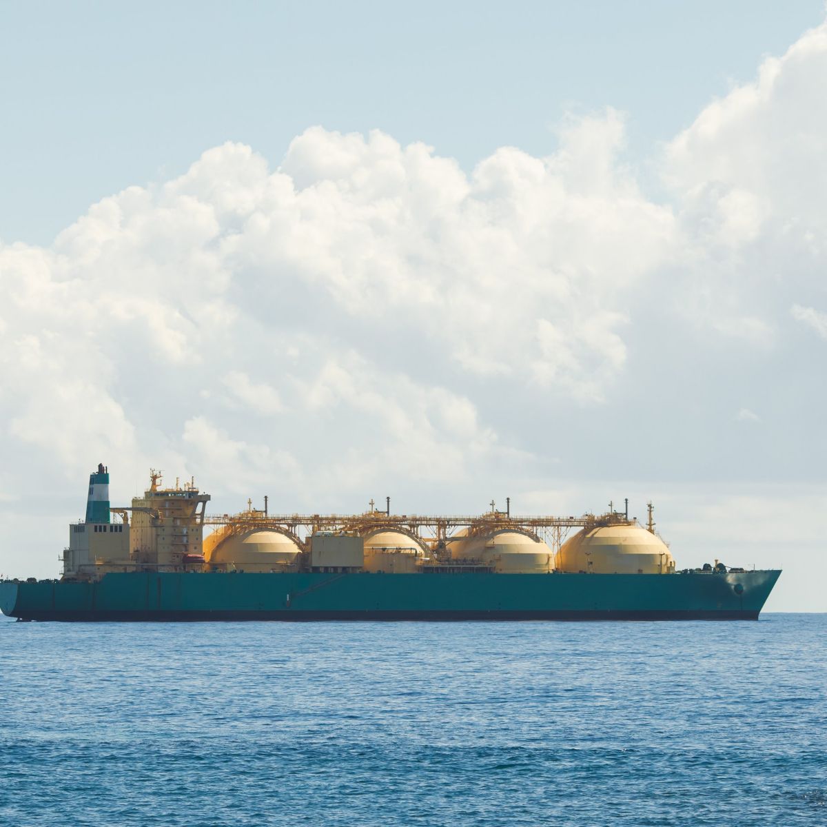 An industrial liquefied natural gas tanker ship cruises across a calm blue ocean under a bright, cloud-filled sky.