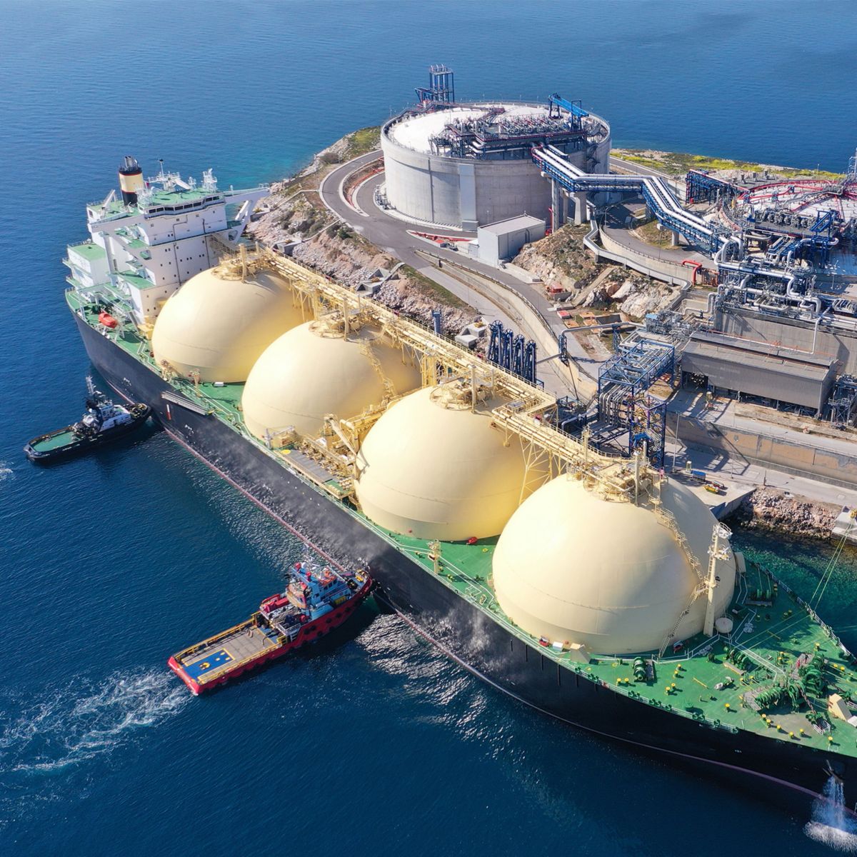 Aerial view of an LNG carrier docked at a coastal terminal with storage facilities, industrial infrastructure, and support vessels nearby.