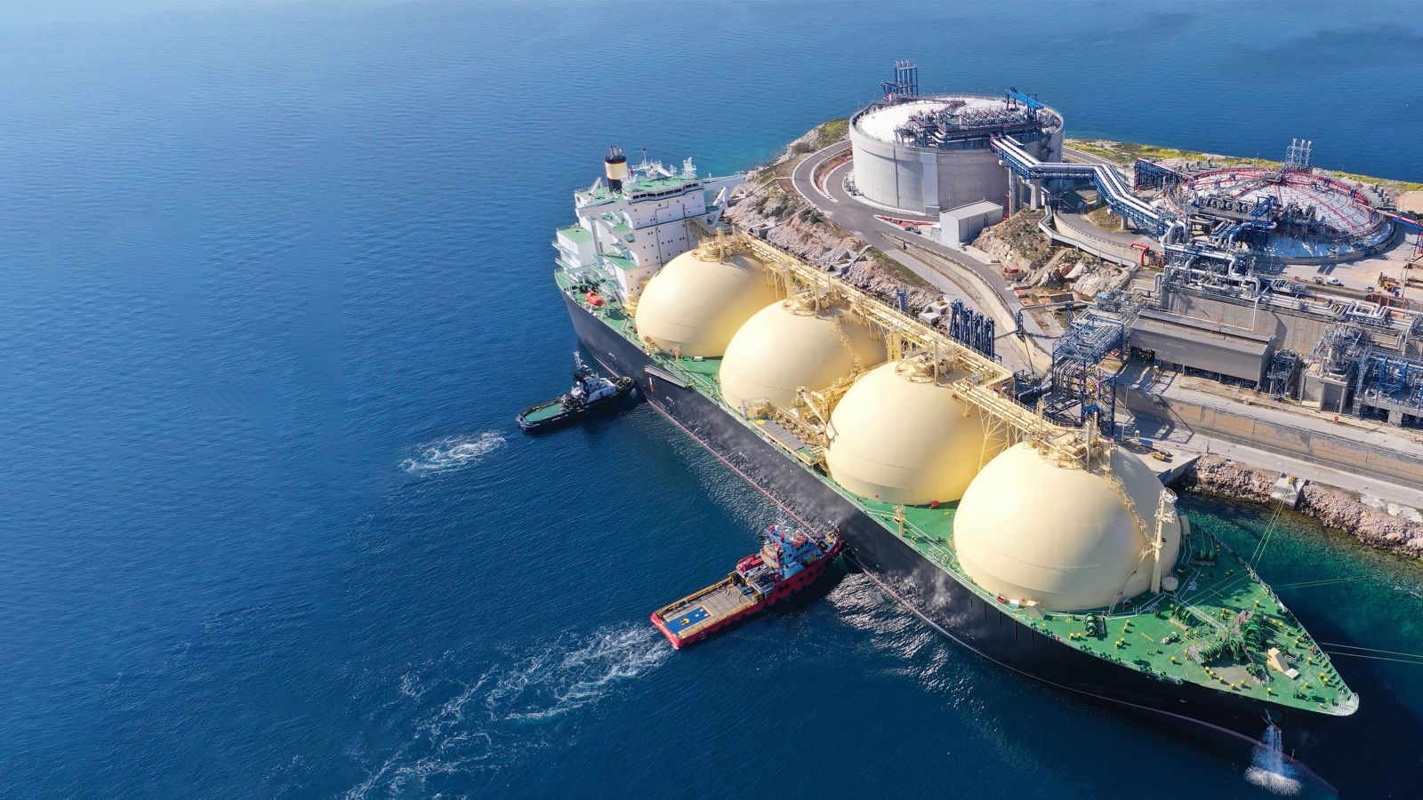 Aerial view of an LNG carrier docked at a coastal terminal with storage facilities, industrial infrastructure, and support vessels nearby.
