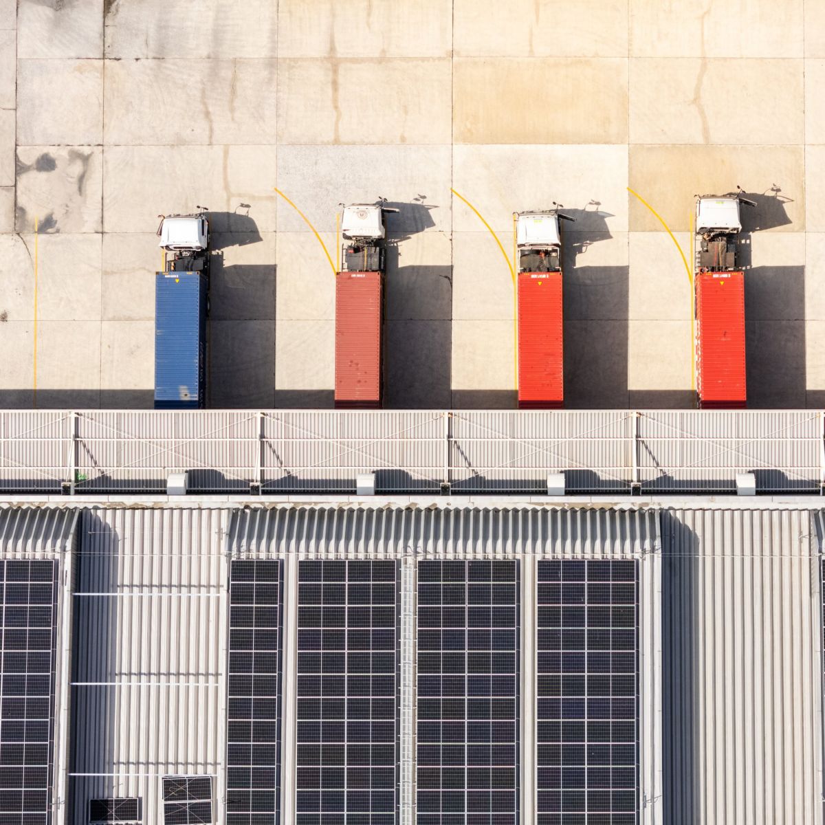 An aerial view displays five semi-trucks parked at a large industrial distribution center featuring a roof covered with solar panels.