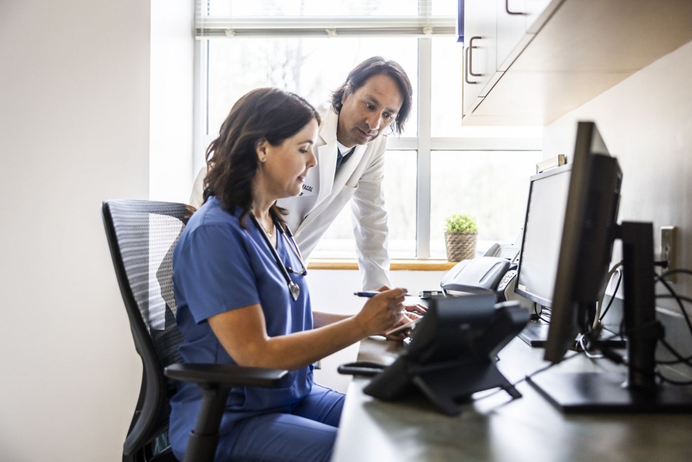 A female nurse and male doctor review digital patient charts together at a workspace.