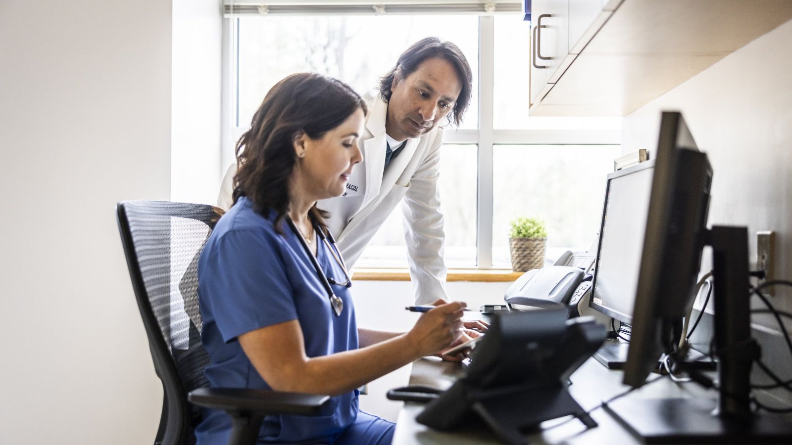 A female nurse and male doctor review digital patient charts together at a workspace.