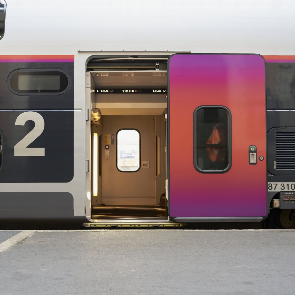A side view of a double-decker passenger train carriage with an open, vibrant purple and orange door at a station platform.