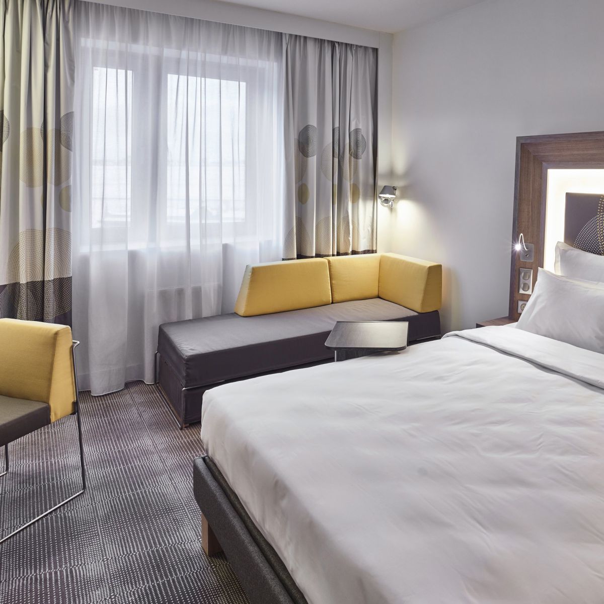 A contemporary hotel suite featuring a plush bed, yellow accents, and natural light.