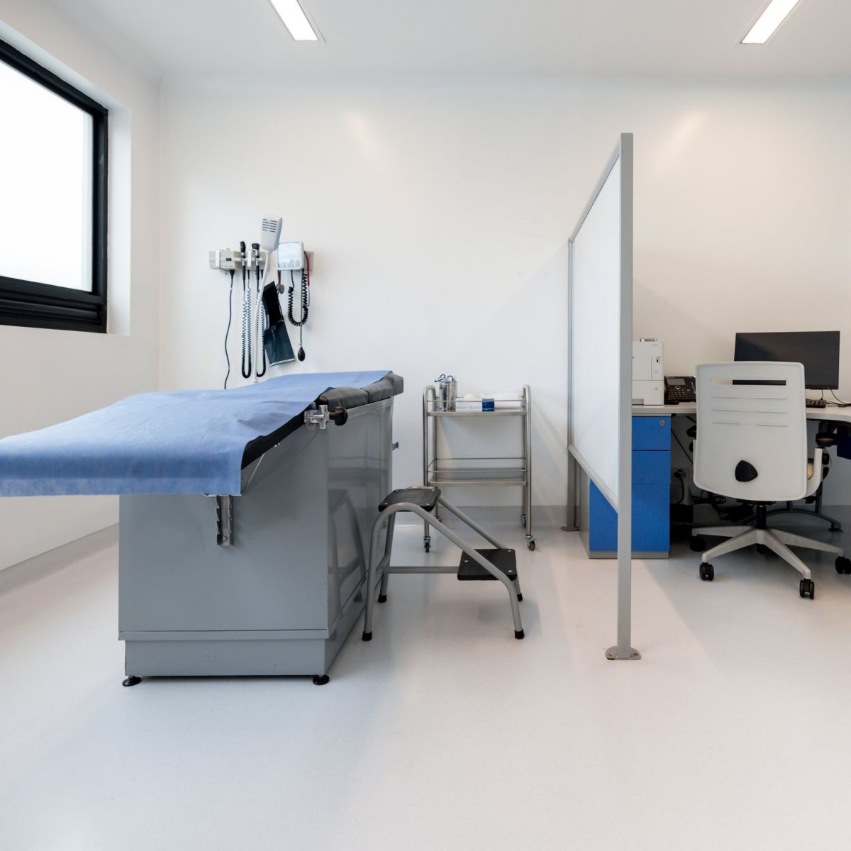 A clean, well-lit medical examination room featuring a patient bed, a divider screen, and a professional workspace area.