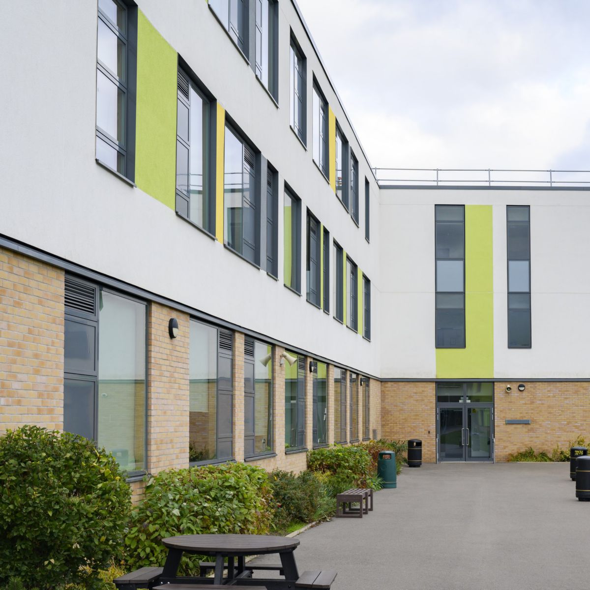 A low-angle shot of a contemporary school building featuring yellow brickwork, white cladding, and vibrant green accents on a cloudy day.