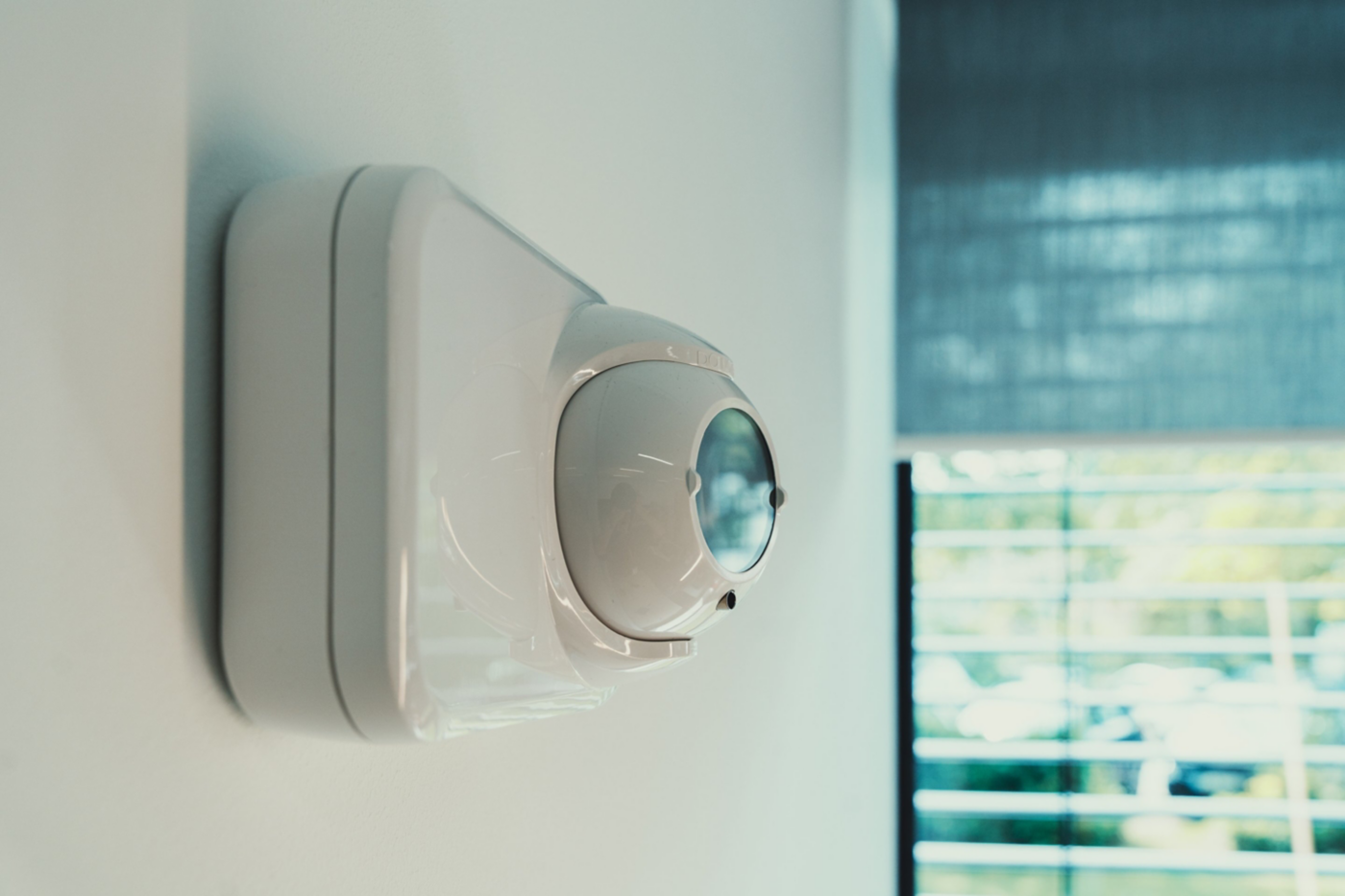 A close-up side profile of a modern white motion detection sensor mounted on a clean office wall near a window.