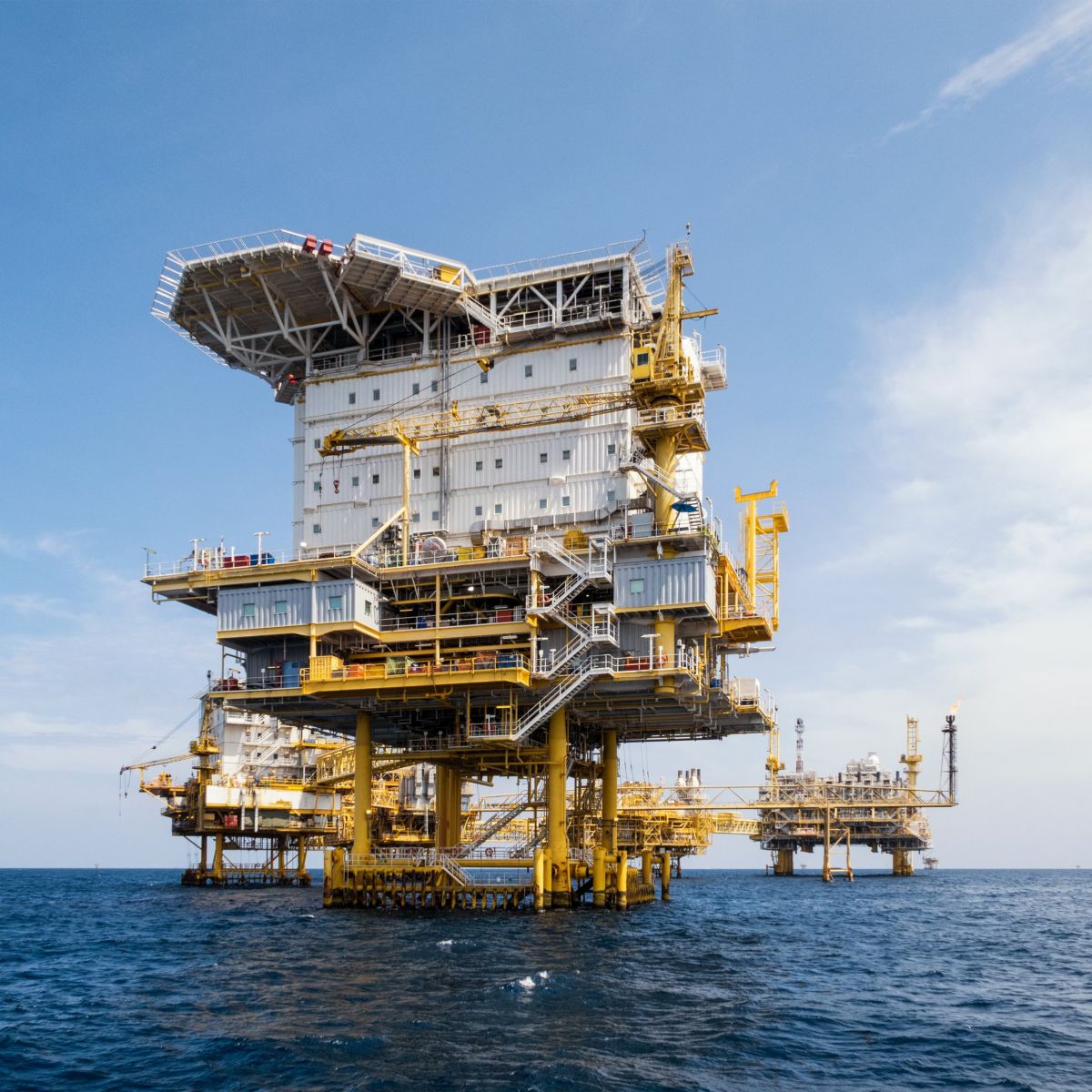 A large industrial offshore oil and gas production platform stands prominently in the middle of the deep blue ocean under a clear sky.
