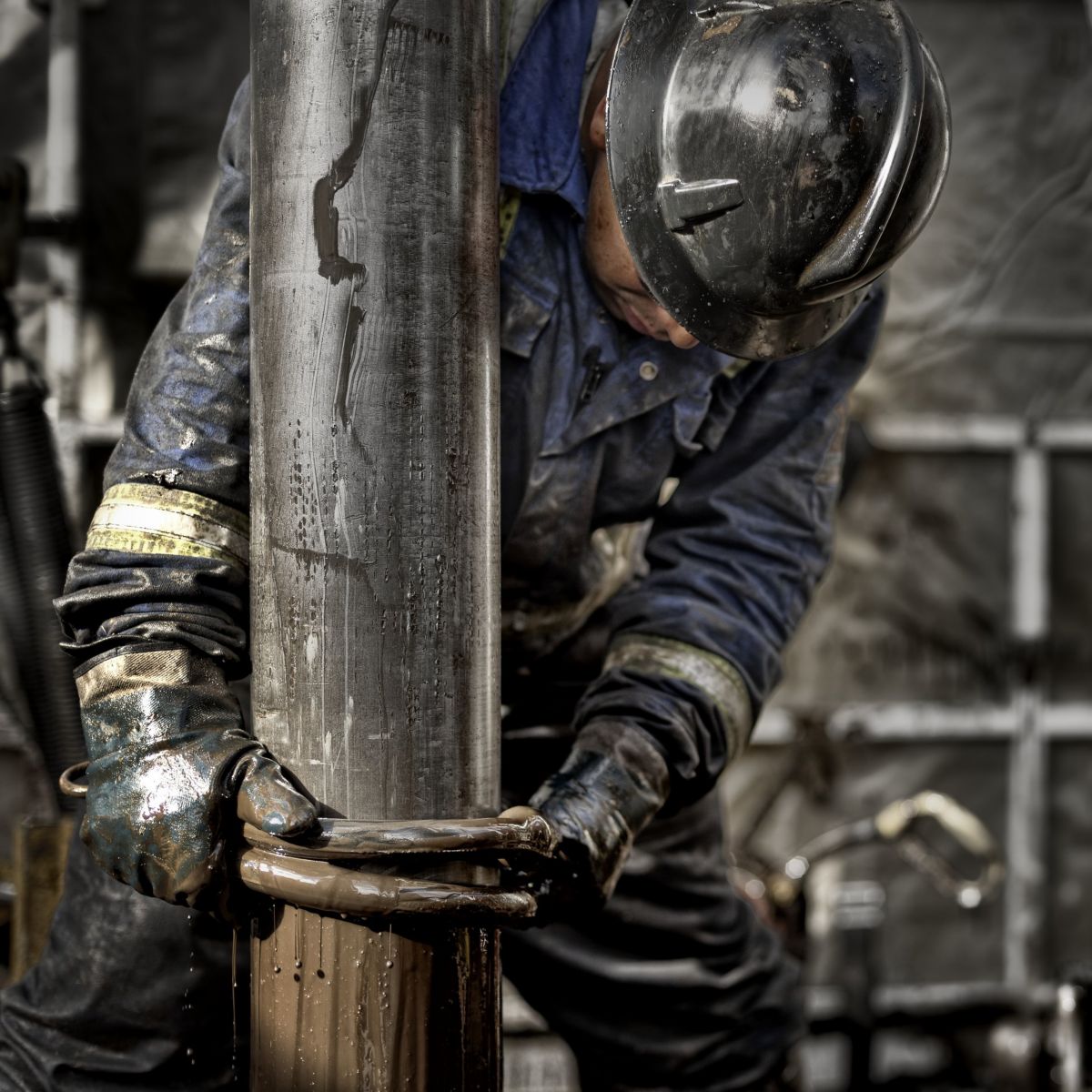 A worker in protective gear manually guides a steel drill pipe on an industrial oil rig.