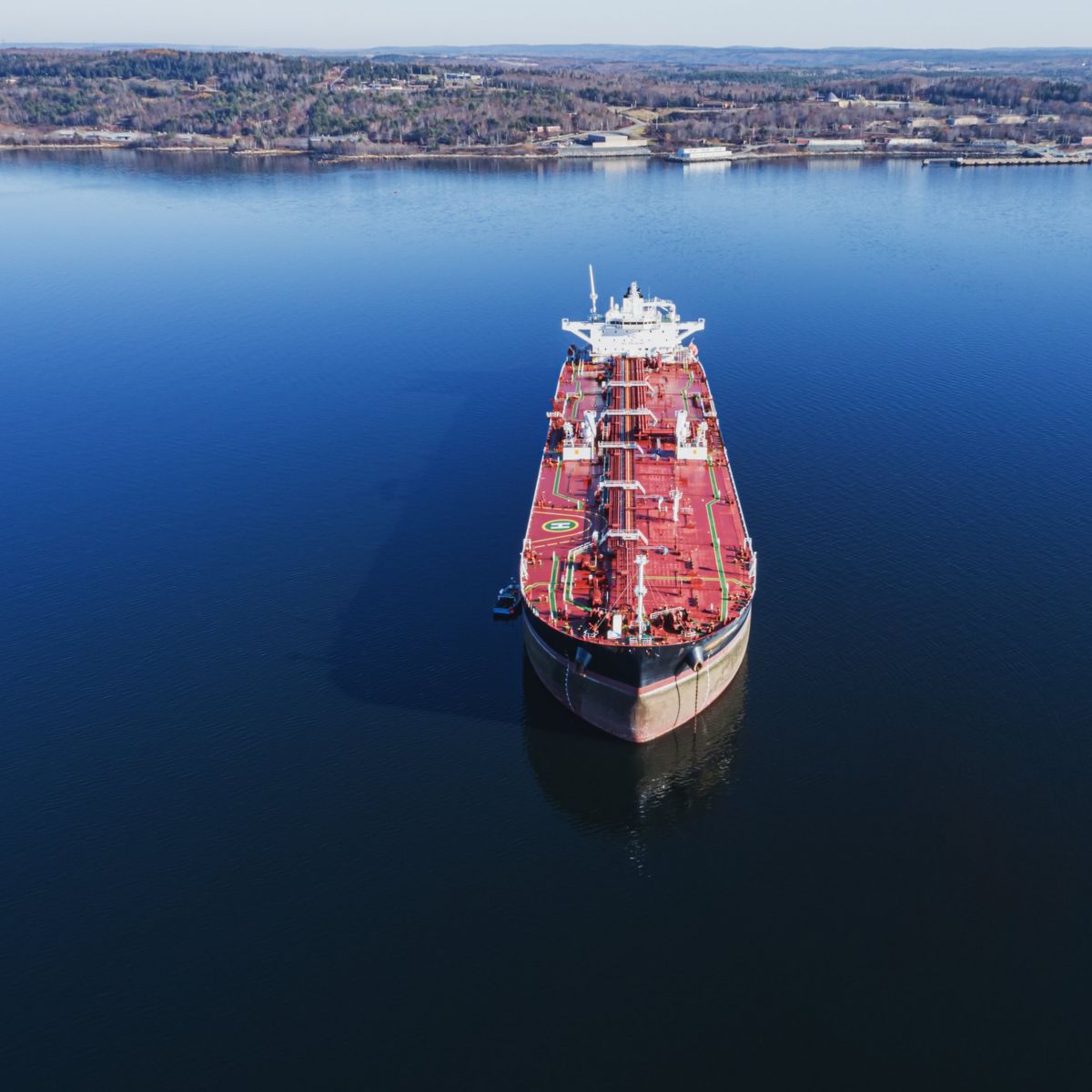 Aerial view of a large red industrial oil tanker anchored in calm, deep blue coastal waters near a forested shoreline.