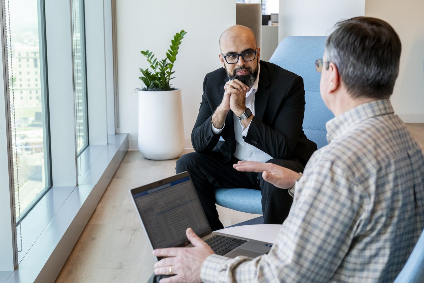 Two people seated facing each other, one using a laptop and the other listening, engaged in a discussion near a window in an office setting.