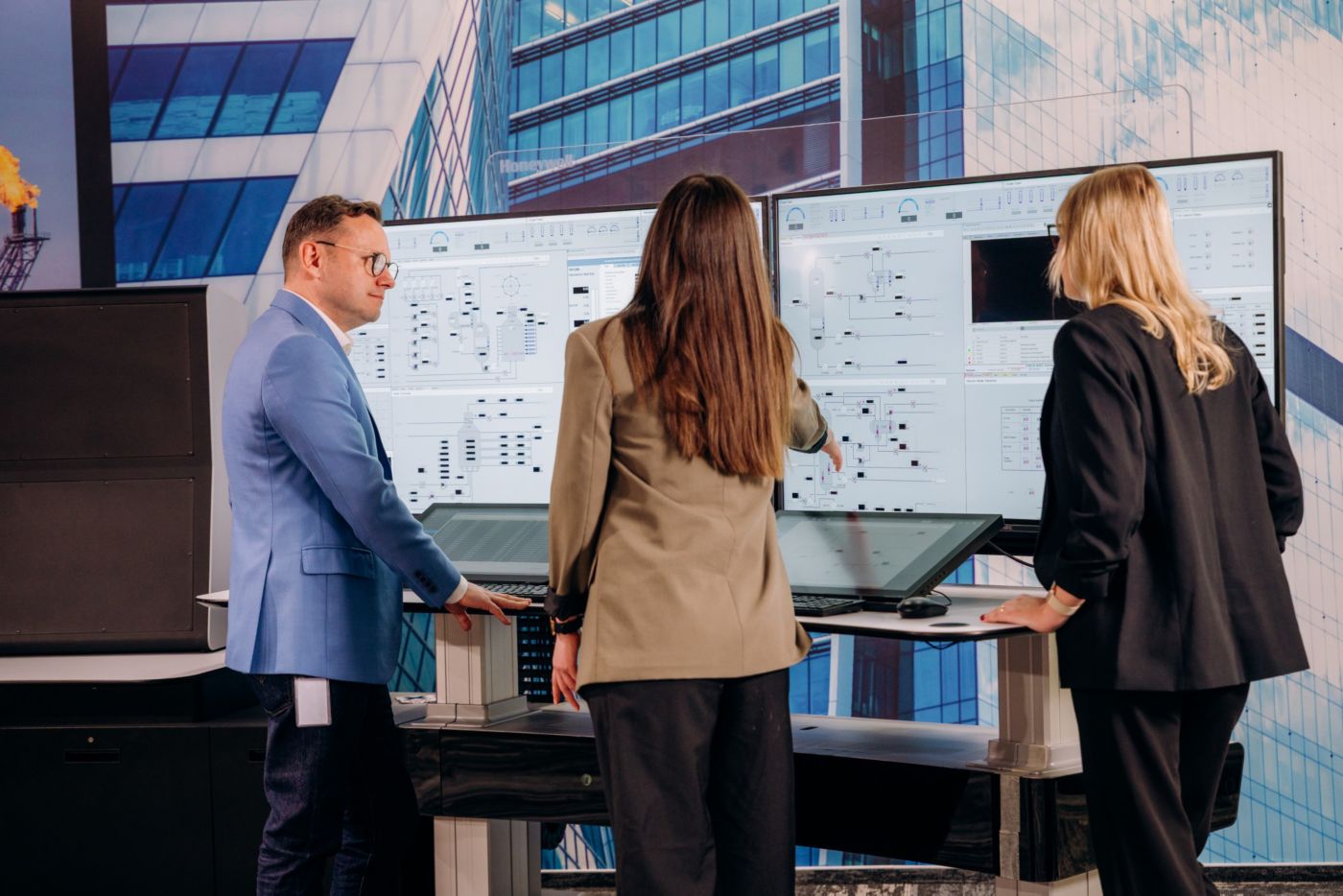 Three people standing at workstation analyzing diagrams and data on multiple large screens, with control interfaces visible in front of them.