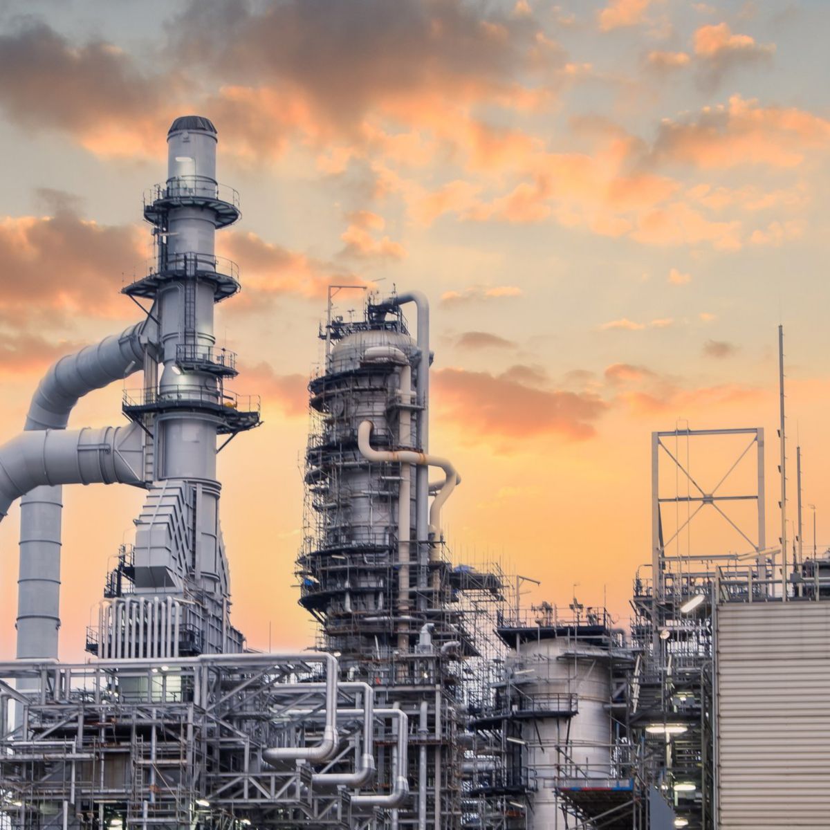 An industrial petrochemical refinery complex silhouetted against a vibrant orange and purple sunset sky.