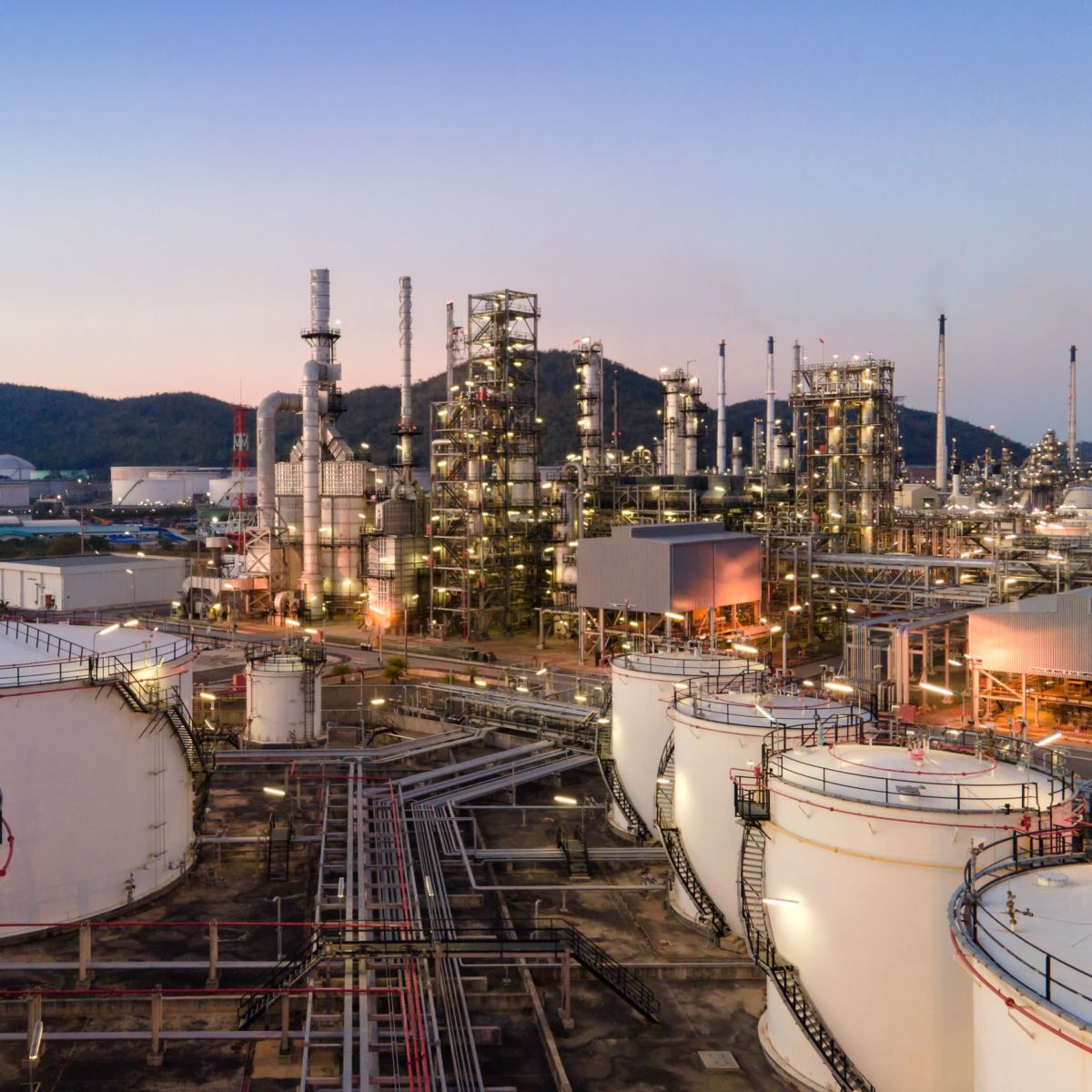 An elevated dusk view of a petrochemical refinery complex featuring illuminated storage tanks and industrial distillation towers against a mountain backdrop.