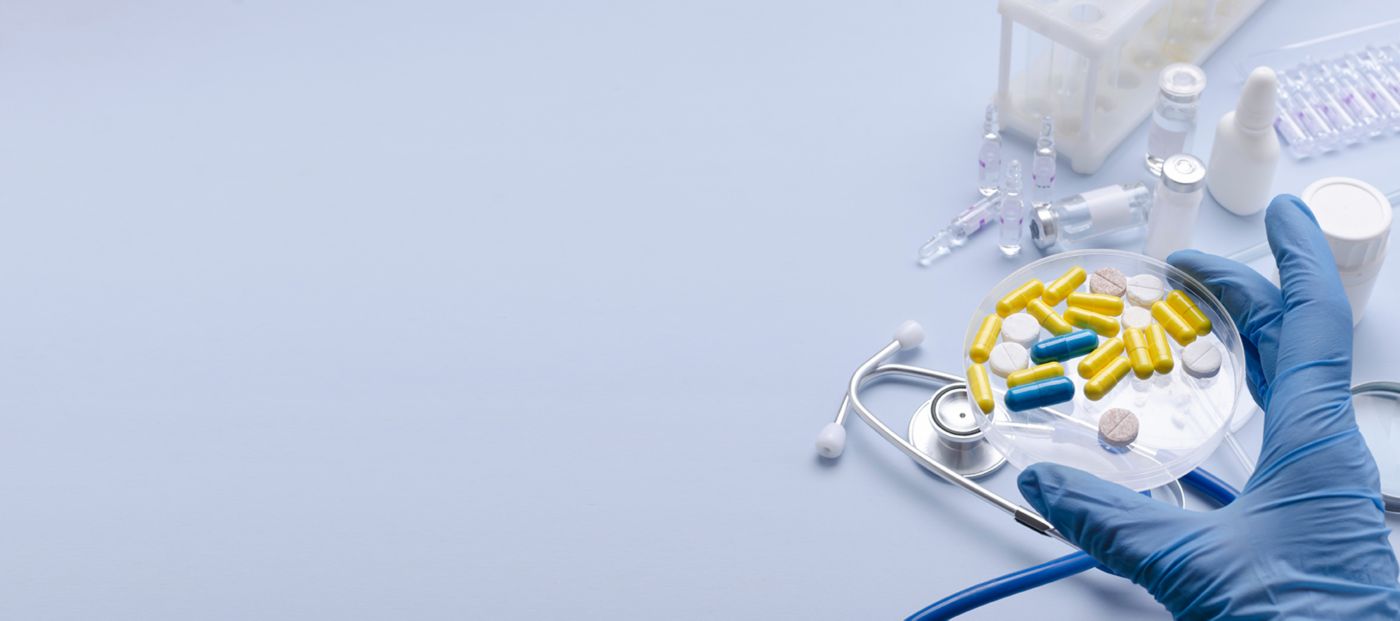 A gloved hand holds a petri dish filled with colorful capsules and tablets beside a stethoscope, vaccine vials, ampoules, and medical supplies in a pharmaceutical laboratory setting. The image represents drug development, medication testing, pharmaceutical research, healthcare innovation, and quality control.