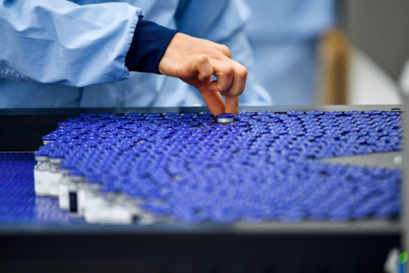 A technician in a cleanroom inspects a line of vaccine vials moving along a conveyor belt for quality assurance.