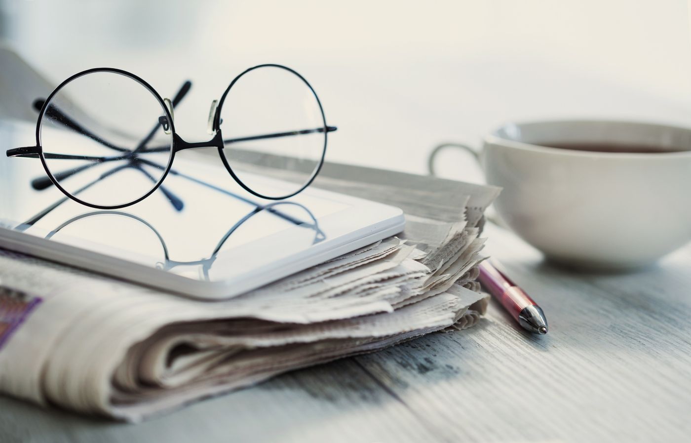 Close-up of glasses resting on a tablet, surrounded by a newspaper, pen, and a cup of coffee.