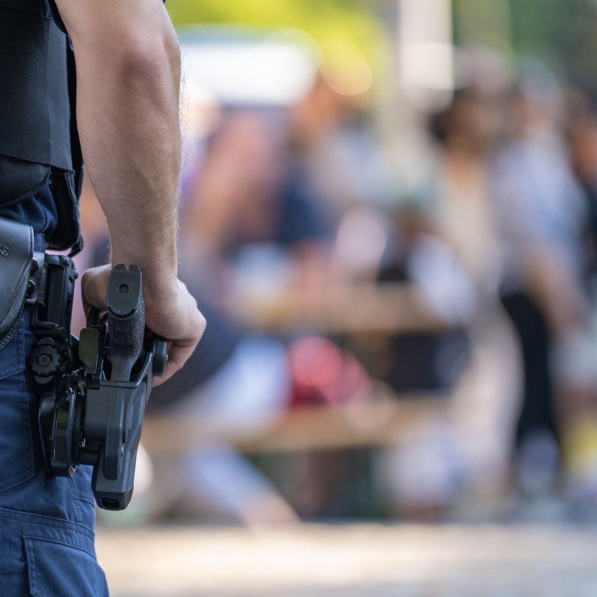 A police officer in tactical gear stands guard with their hand resting on a holstered sidearm against a blurred crowd background.
