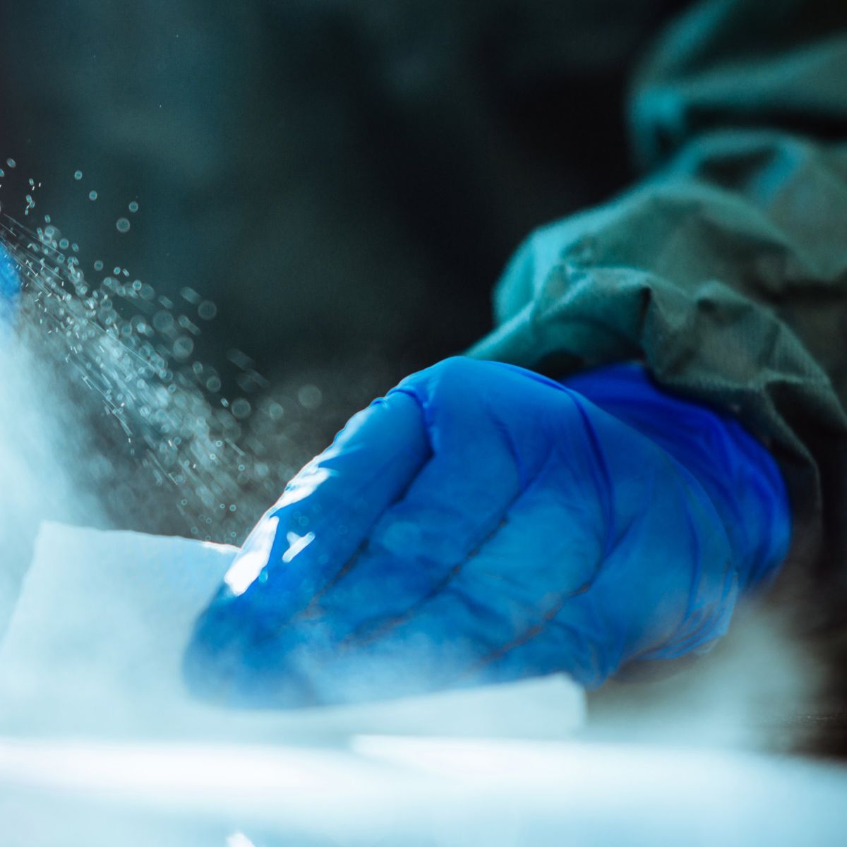 A worker wearing blue nitrile gloves sprays disinfectant onto a surface while cleaning it with a cloth in a sterile industrial environment.